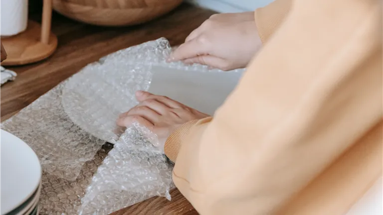 A woman carefully wraps dishware in bubble wrap on a wooden kitchen counter.