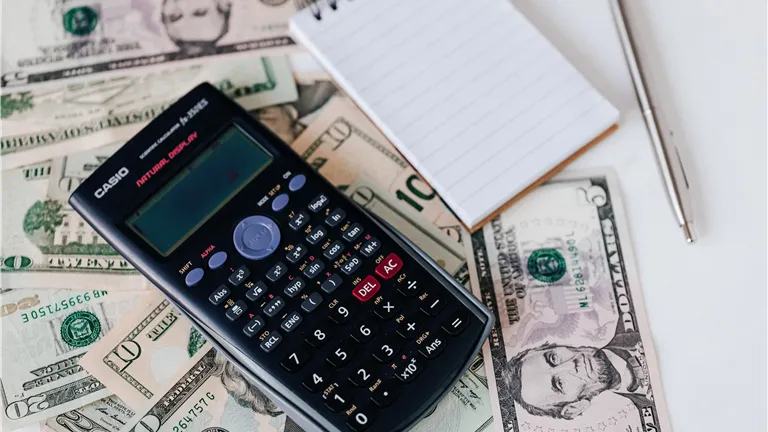 From above electronic calculator and notepad placed over United States dollar bills together with metallic pen for budget planning and calculation