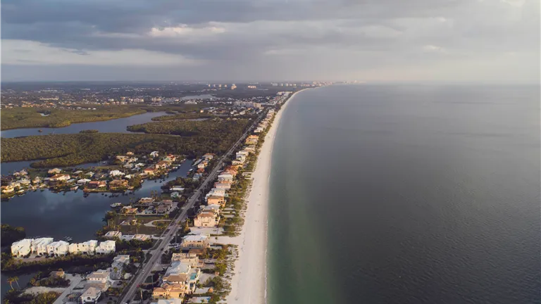Stunning aerial view of coastal homes lining a serene beach with the ocean and clouds in the distance.