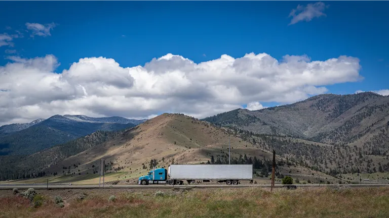 A blue semi-truck travels along a scenic highway with mountains and cloudy skies in the background.