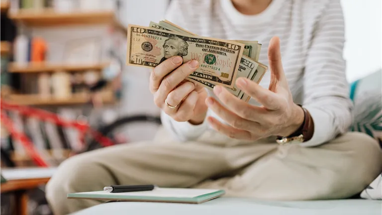 Man wearing casual clothes counts dollar bills while sitting on a sofa indoors.